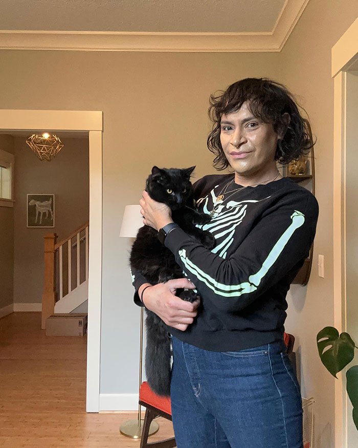 Person wearing a skeleton print sweater holding a black cat indoors, related to trans chef of the year media coverage.