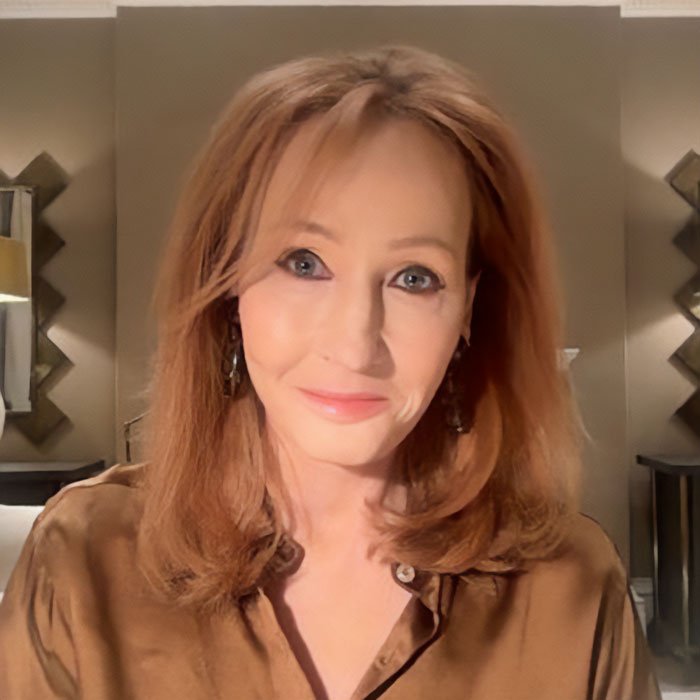 J.K. Rowling wearing a brown blouse, smiling softly in a warmly lit indoor setting with decorative mirrors in the background