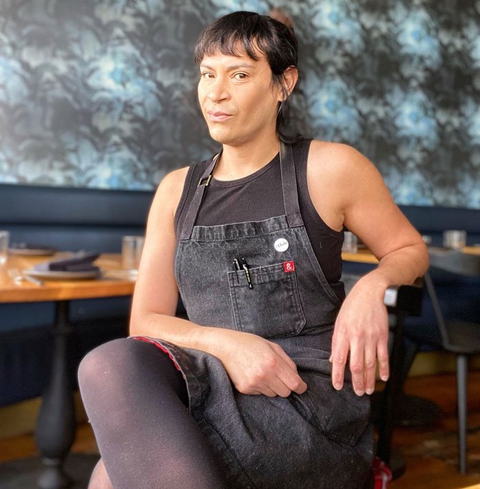 Trans chef of the year sitting in a restaurant wearing a black apron and sleeveless shirt, posing confidently.