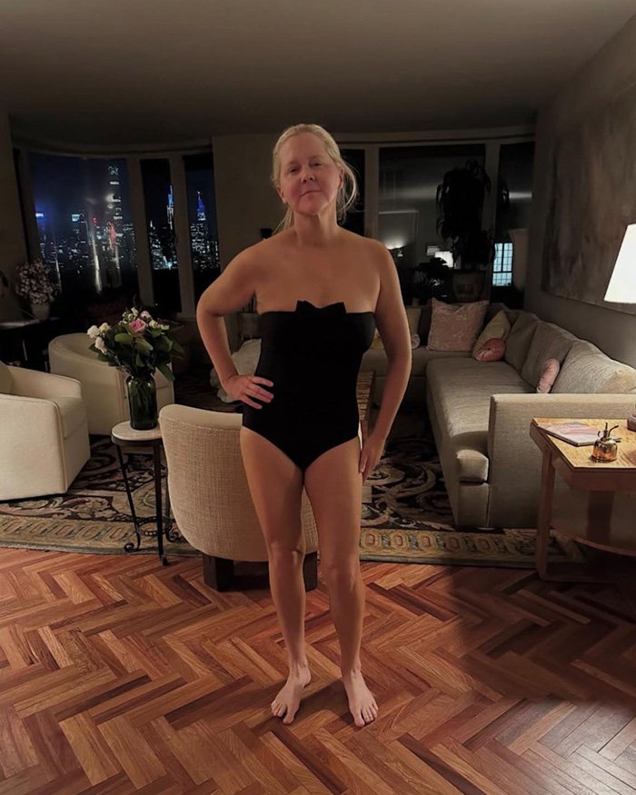 Amy Schumer in a black swimsuit standing in a living room with city lights visible through the windows at night.