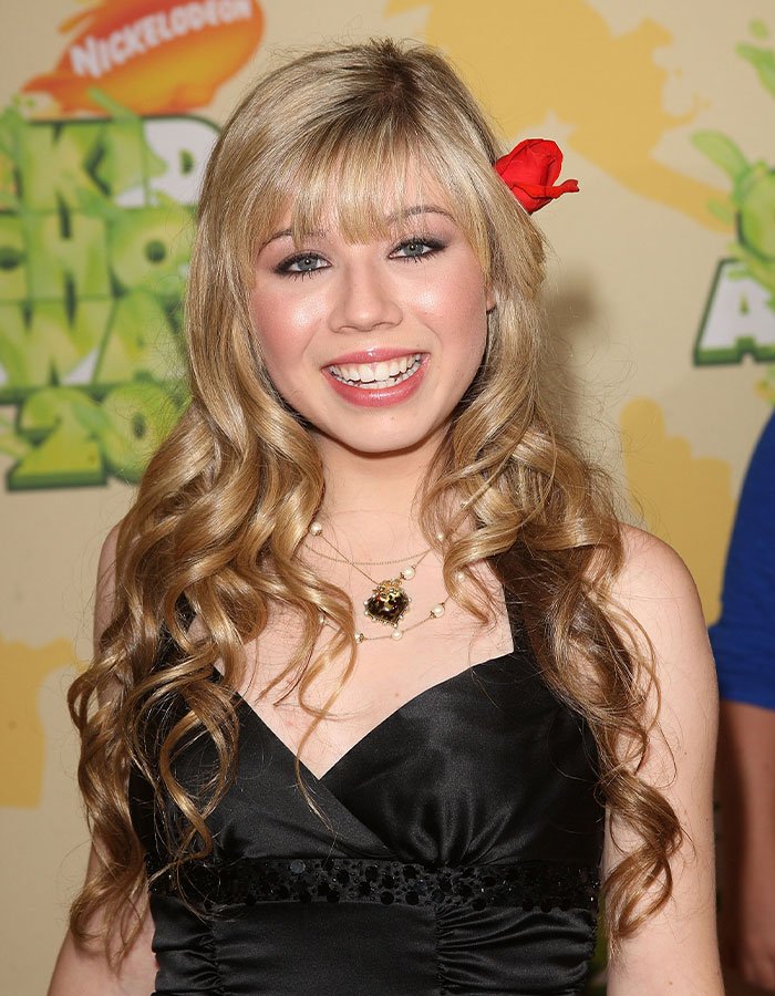 Jennette McCurdy smiling at Nickelodeon event, with long curly blonde hair and a red flower in her hair.