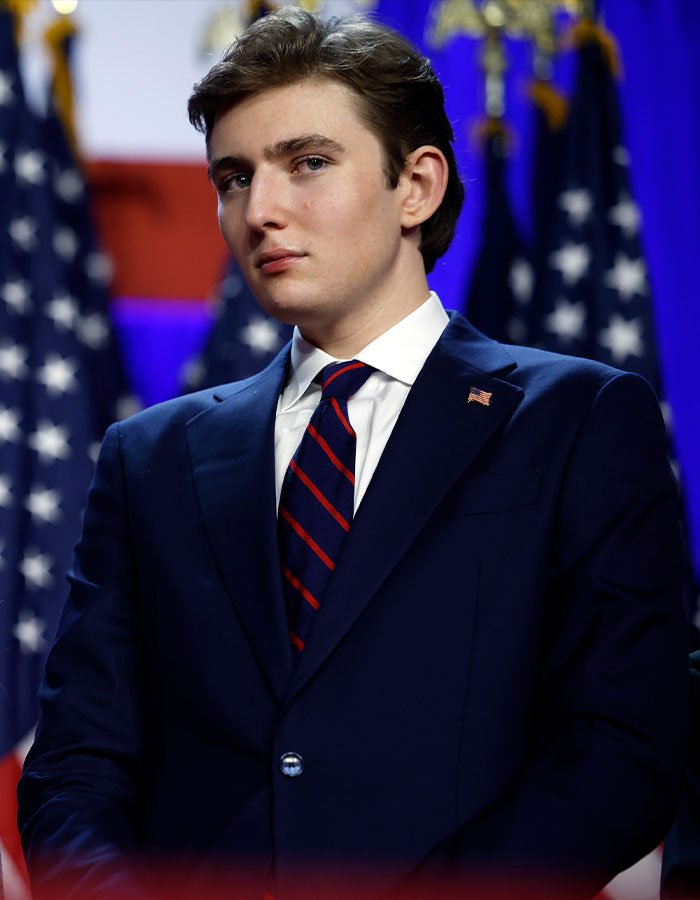 Barron Trump in a navy suit and striped tie, standing in front of American flags at a formal event.