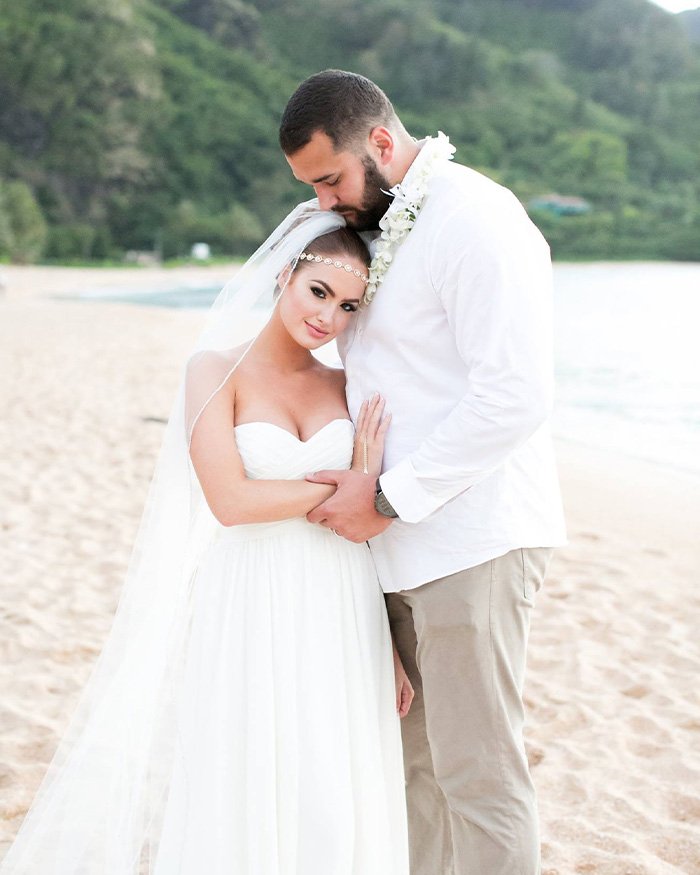 Bride in white dress and veil embraces groom wearing white shirt and lei on a beach, related to former NFL star's ex-wife news.