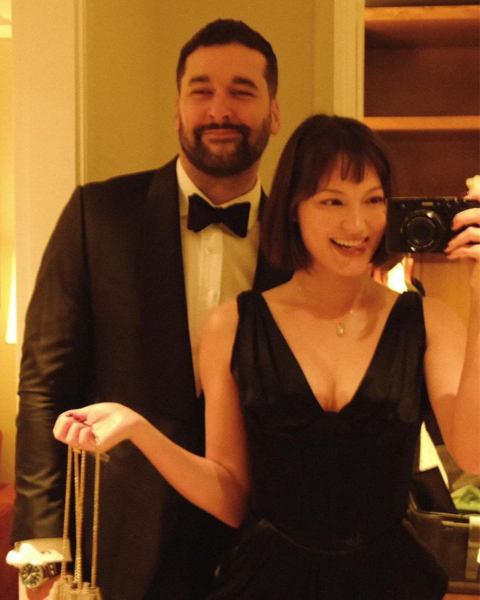 Former NFL star's ex-wife takes selfie in black dress while man in tuxedo stands behind her, smiling at camera.