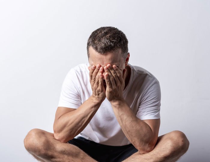 Man in a white shirt sitting cross-legged with face buried in hands, conveying distress and unemployment emotions.