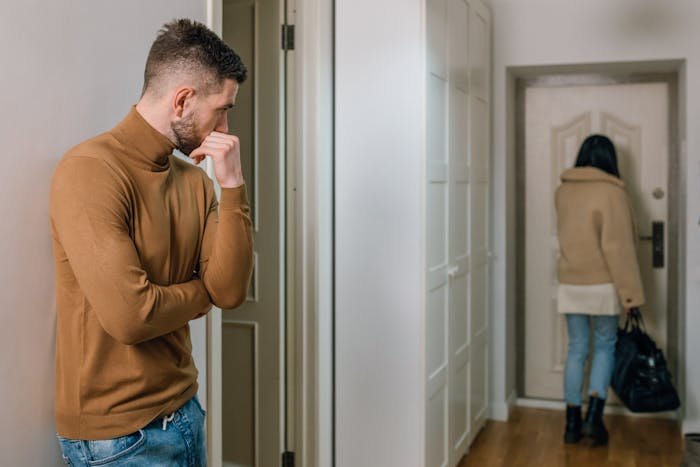 Man in a brown sweater looking thoughtful as an unemployed woman leaves a room carrying a black bag.