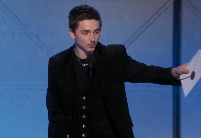 Actor Timothée Chalamet in a black suit speaking at a podium during the Golden Globes, sparking media buzz about snubs.