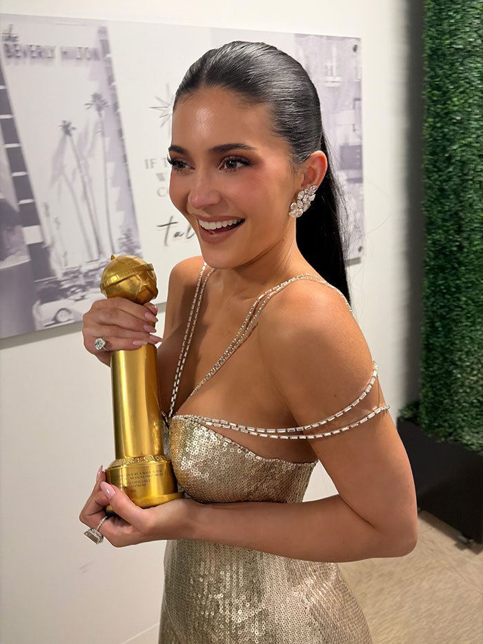 Woman in a gold dress smiling and holding a Golden Globe award, related to Timothée Chalamet Golden Globes post news.