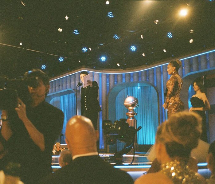 Timothée Chalamet on stage at the Golden Globes with a golden trophy, spotlight, and attendees in the audience.