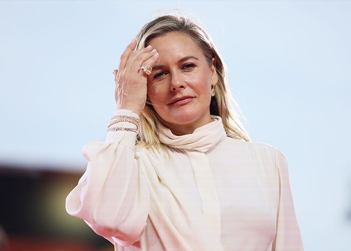 Alicia Silverstone in a light-colored outfit at an event, touching her hair with a neutral expression.