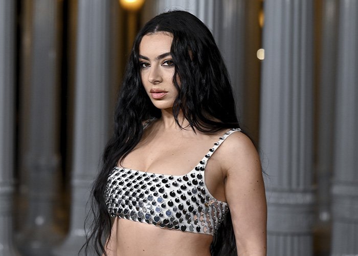 Charli XCX with long black hair wearing a silver studded braless top at an elegant columned venue.