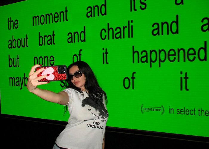 Charli XCX wearing sunglasses and a graphic tee taking a selfie in front of a large green screen with black text.