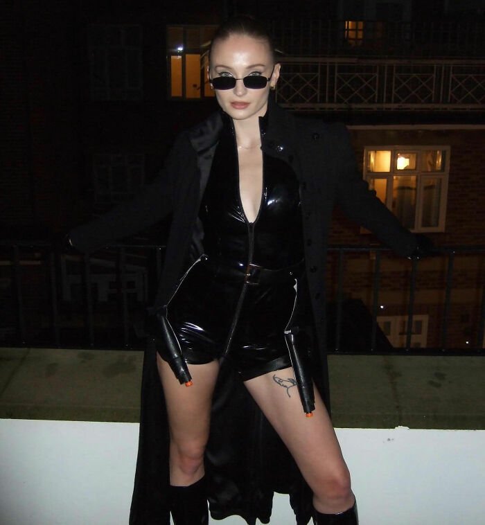 Sophie Turner in a black outfit and sunglasses showcasing her ripped Lara Croft transformation for Tomb Raider at night.
