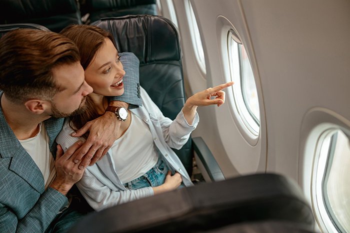 Couple seated on a plane, woman points out the window while man looks on, illustrating guy’s heroic actions theme.