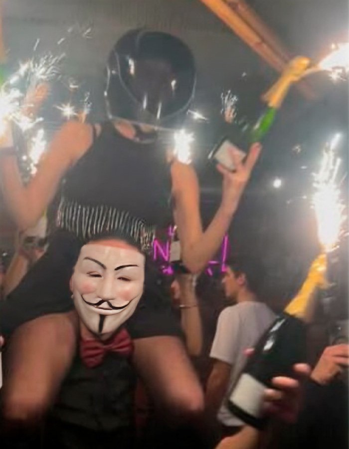 Party scene with a waitress wearing a helmet and celebrating, holding sparklers and champagne bottles in a lively bar environment.