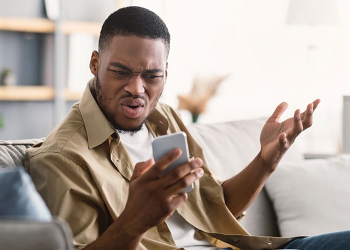 Man sitting on couch looking frustrated at phone, illustrating open relationship plan backfiring with younger coworker.