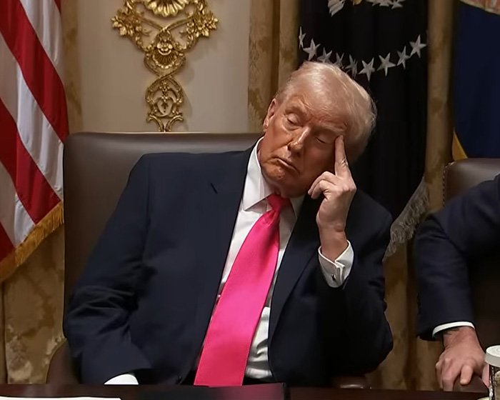 Donald Trump in a suit with a droopy face, resting his head on his hand during a formal meeting in a government room.