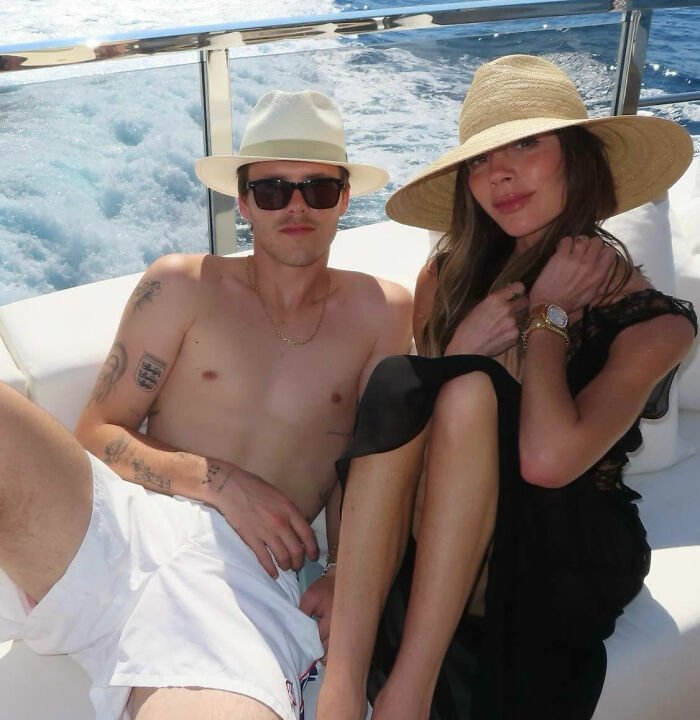 Cruz Beckham shirtless in white hat and shorts relaxing with a woman in a wide-brimmed hat on a boat at sea.