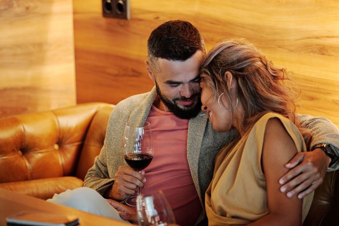 Couple sharing intimate moment on a leather couch, woman smiling while man holds a glass of wine in cozy setting