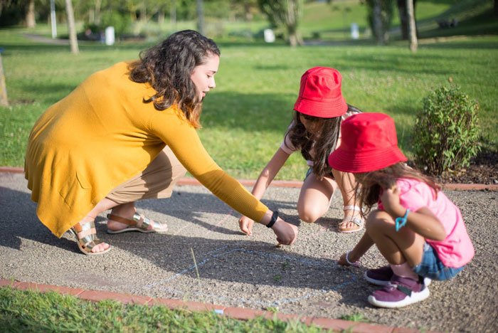 Woman refusing to pay for friends babysitter, sitting outdoors playing with children wearing red hats on a sunny day.