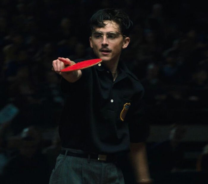 Young man playing table tennis intensely, showcasing focus and skill in a dimly lit indoor setting for Oscar nominations.