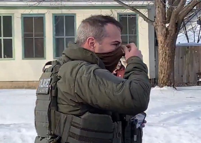 Law enforcement officer wearing tactical gear and adjusting face covering outside a snow-covered residential area.