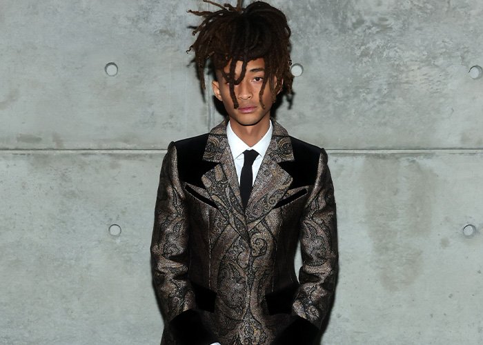 Jaden Smith wearing a patterned suit and tie posing against a concrete wall, linked to Black Brooklyn Beckham and Louboutin.