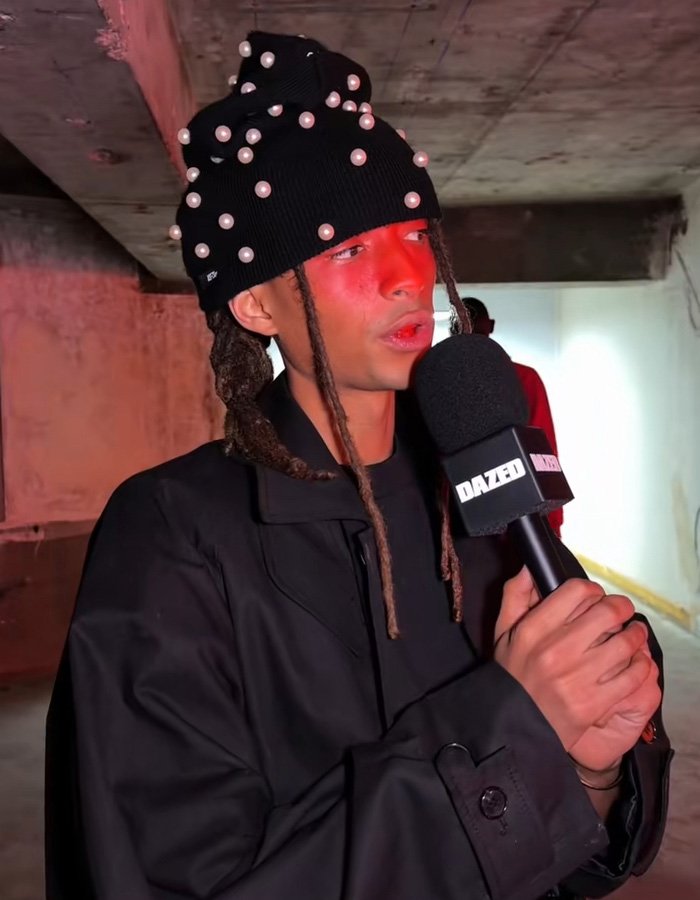 Jaden Smith in a black pearl-studded hat and coat speaking into a DAZED microphone during an interview.