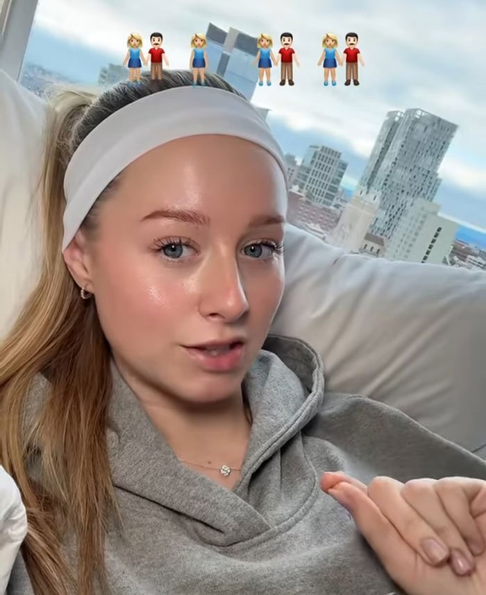 Young woman sharing story about having 7 parents, seated indoors with city skyline visible through window behind her.
