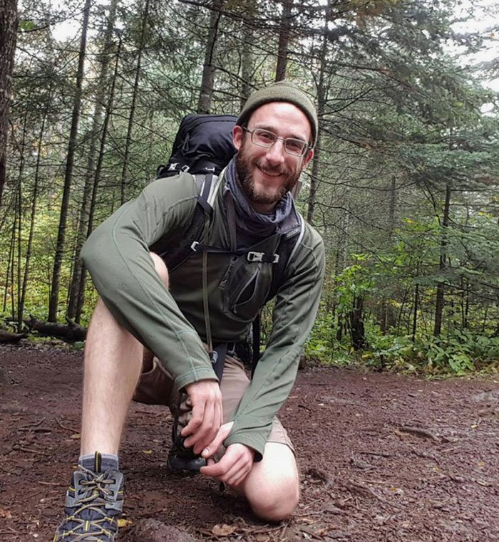 Man wearing glasses and hiking gear kneeling on a forest trail reflecting the story about Alex Pretti’s fate and government care.