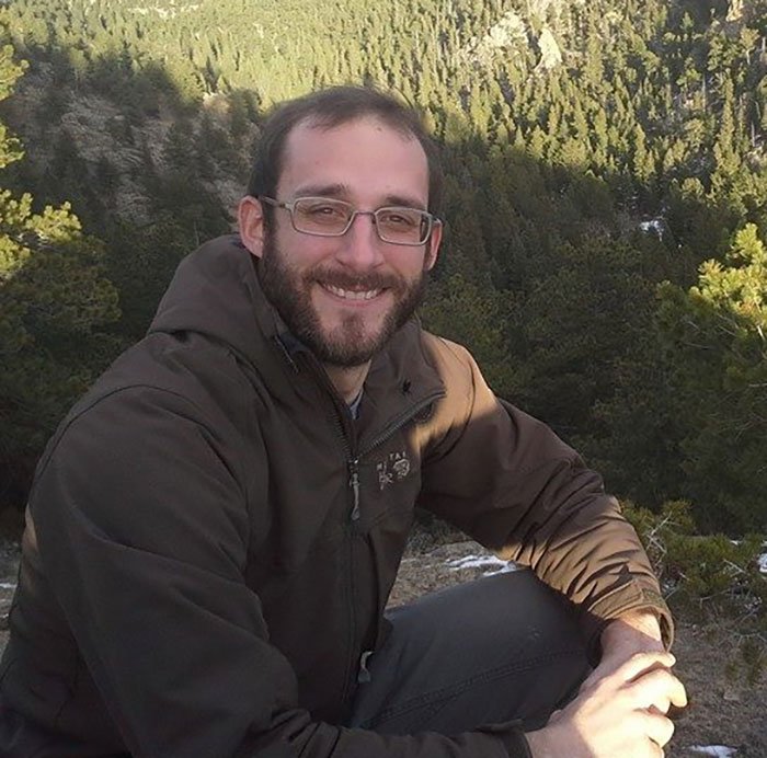 Man wearing glasses and a brown jacket sitting outdoors in front of a forest, relating to Alex Pretti government care story.
