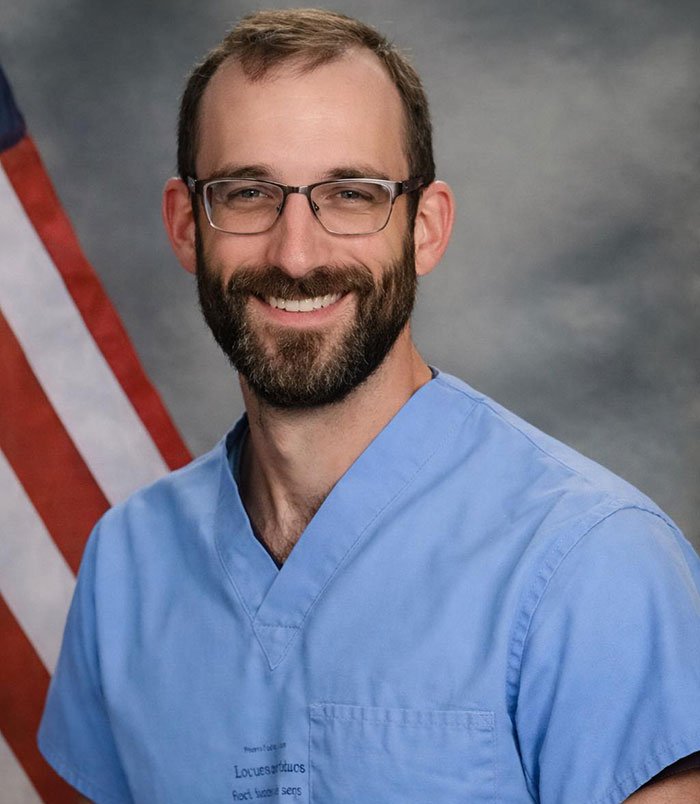 Man with glasses and beard wearing blue scrubs, smiling in front of an American flag, related to Alex Pretti’s parents learning his fate.