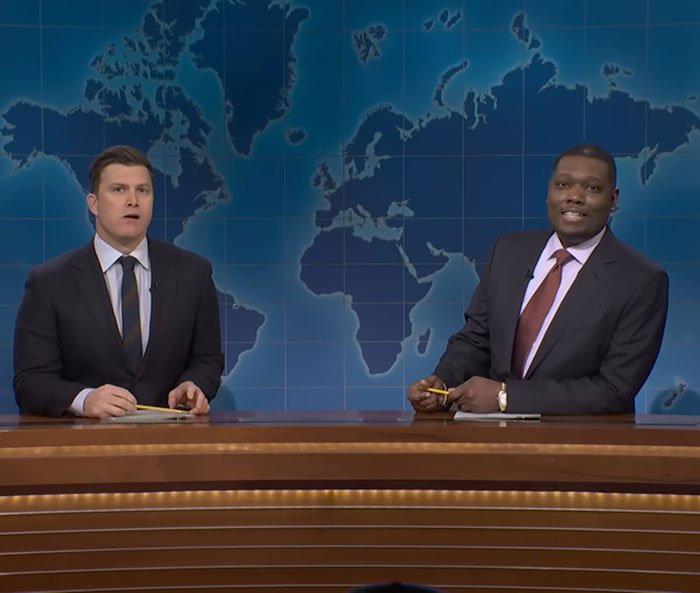 Two Weekend Update anchors on SNL set with world map backdrop, discussing a joke about ICE sparking debate.