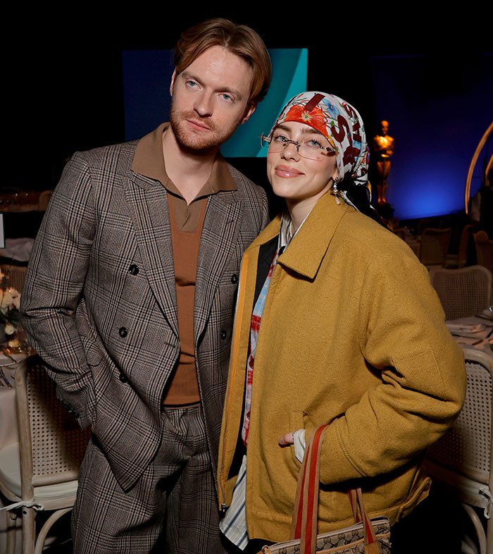 Billie Eilish wearing a headscarf and glasses posing with a man in plaid suit, reflecting the American way of thinking.