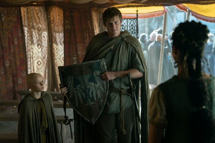 A medieval scene with two characters in cloaks, one holding a shield, comparing Tanzyn Crawford's height to Tansell Too-Tall.