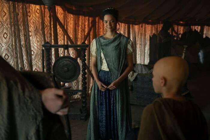 Young woman in medieval attire standing indoors with patterned curtains, related to Tanzyn Crawford's height compared to Tansell Too-Tall.