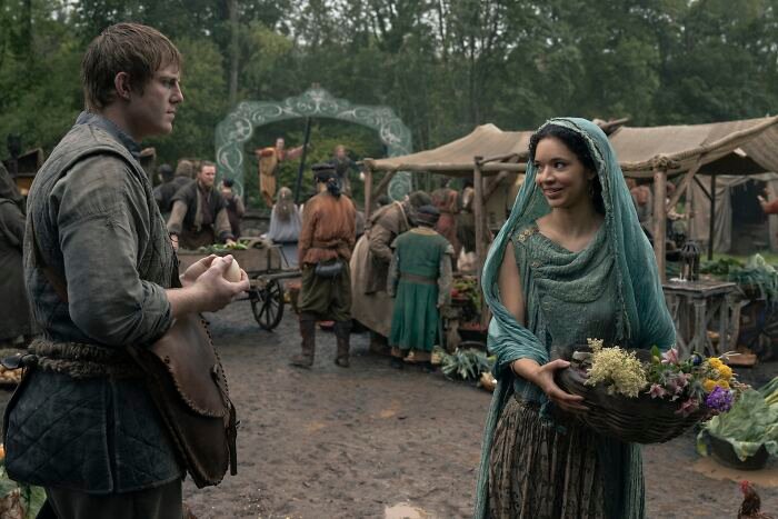 Two people dressed in medieval clothing in a market scene, discussing Tanzyn Crawford height compared to Tansell Too-Tall.