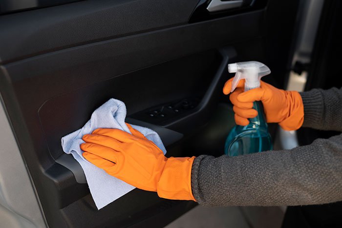 Hands wearing orange gloves cleaning inside of car door with cloth and spray bottle, related to woman gives birth in friends car.