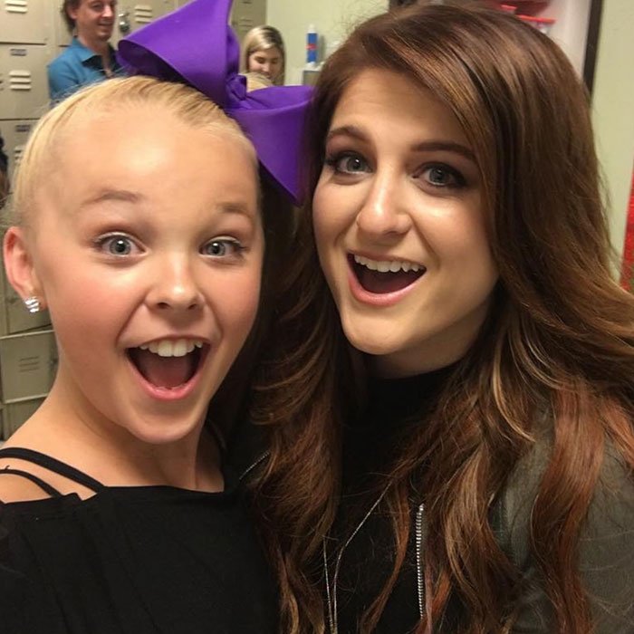 JoJo Siwa with a large purple bow and another woman smiling excitedly in a casual indoor setting.