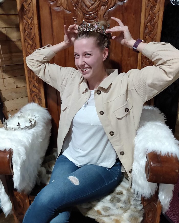 Woman smiling and placing a crown on her head while sitting on an ornate wooden throne chair indoors