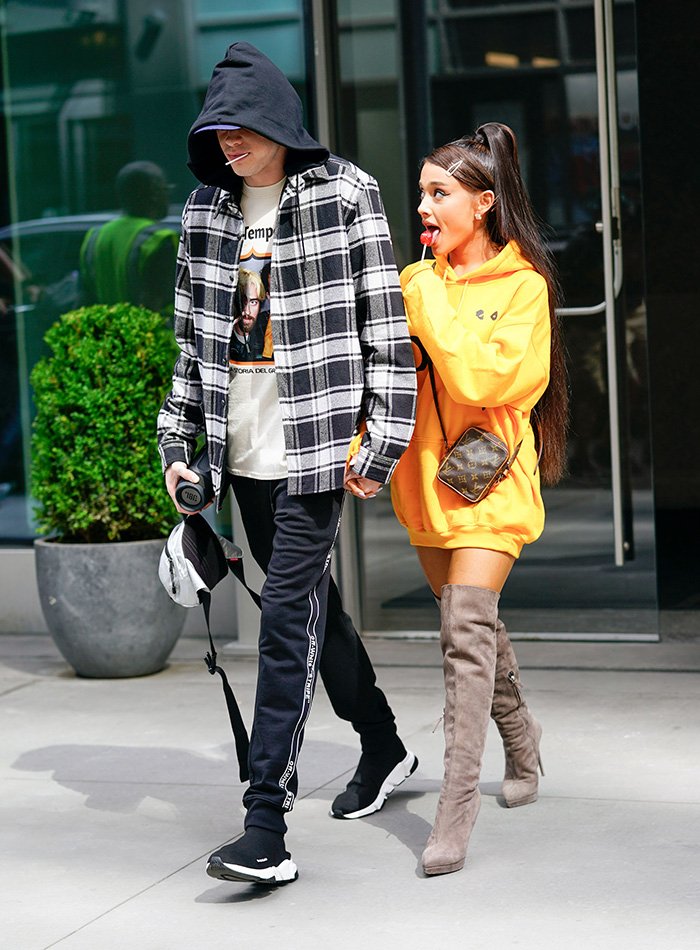 Ariana Grande wearing thigh-high boots and an oversized sweatshirt walking outside with a companion on a sunny day.