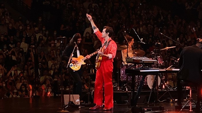 Harry Styles performing live on stage in a red outfit with a large audience during his new tour concert.