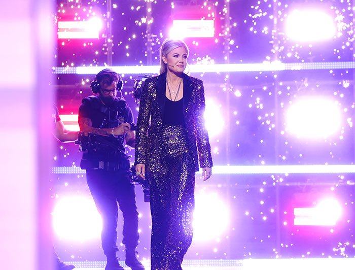 Erika Kirk walking on stage in a glittery suit with bright lights and sparks creating a dramatic atmosphere amid backlash.
