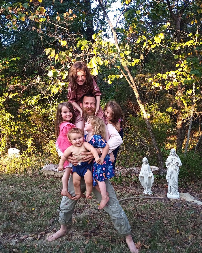 Man living off the grid with children near religious statues in a wooded poverty setting after arrest.