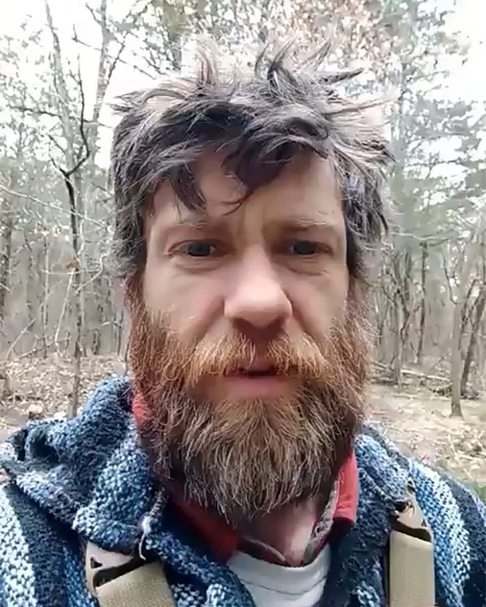Man with unkempt beard and hair wearing a striped hoodie in a forest, symbolizing poverty and off the grid living.
