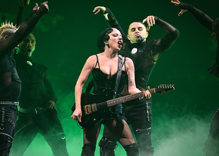 Lady Gaga performing on stage with guitar and dancers in dark outfits during bold ICE speech at her concert.