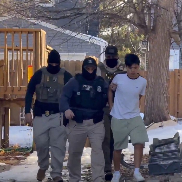 ICE officers escorting a young man in a white shirt during an enforcement operation in a snowy residential area.