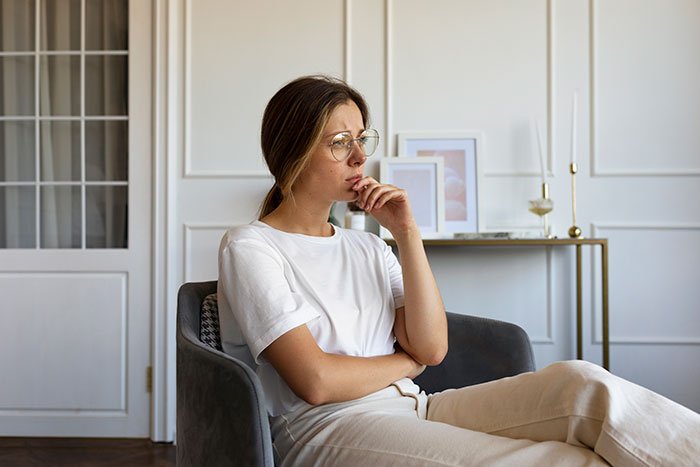 Young woman thoughtfully sitting in chair, symbolizing mom against daughter getting a DNA test, reflecting family conflict and curiosity.