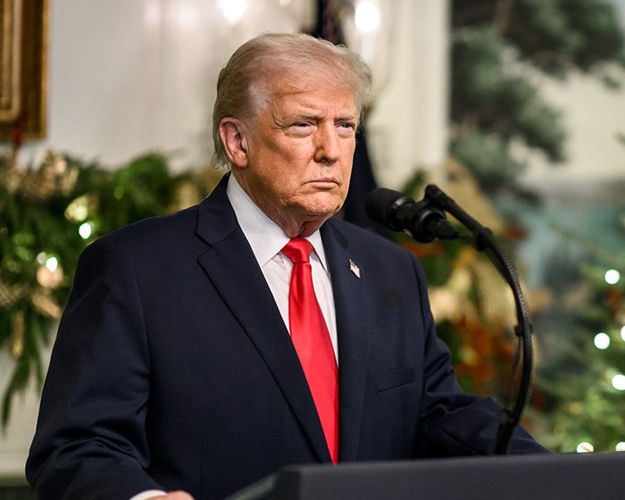 Former president Trump speaking at a podium during a formal event with holiday decorations in the background.