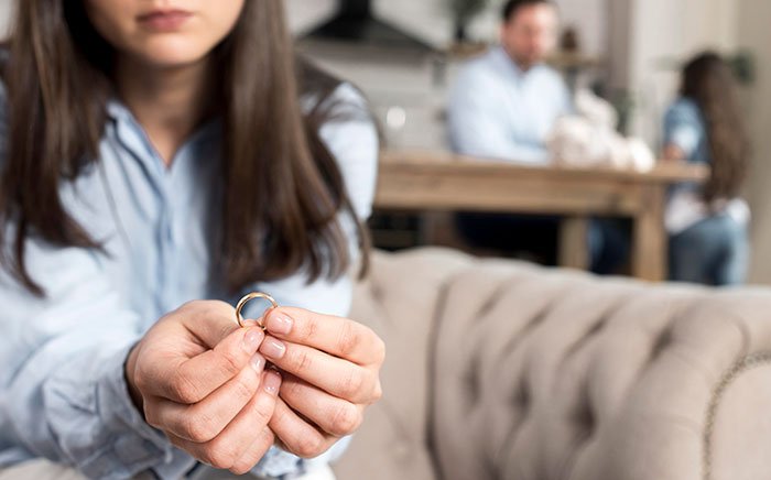 Woman holding a wedding ring looking upset while a man and another woman argue in the background at home.